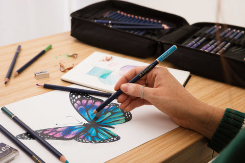 A close up of an artist's hand holding a Castle Gold coloured pencil. She is using it to colour in a butterfly drawing.